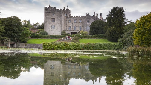 Historic stone castle at Sizergh reflected in a calm pond, surrounded by greenery under a cloudy sky.
